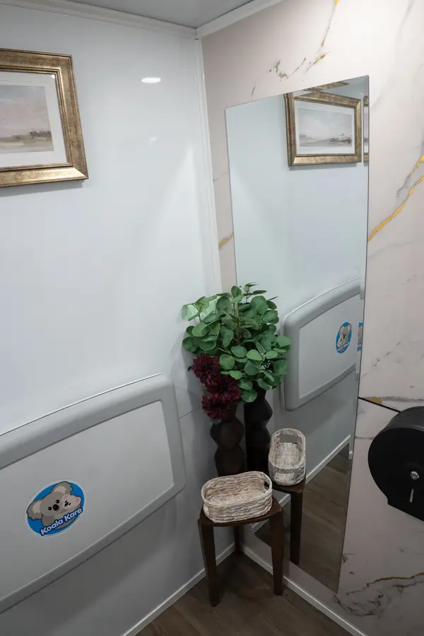 Corner of the women's suite with gold-framed art, a full-length mirror, greenery, and a basket on a wooden stool
