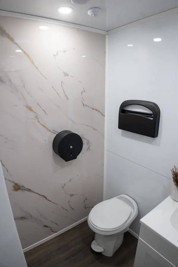 Toilet with a black tissue dispenser on a marble and gold wall, a seat cover dispenser, and recessed ceiling lights
